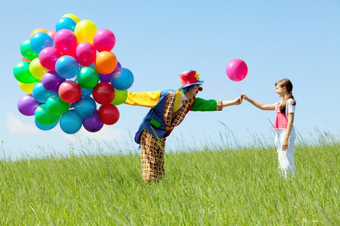 Clown with balloon bouquet giving one to a girl in a field